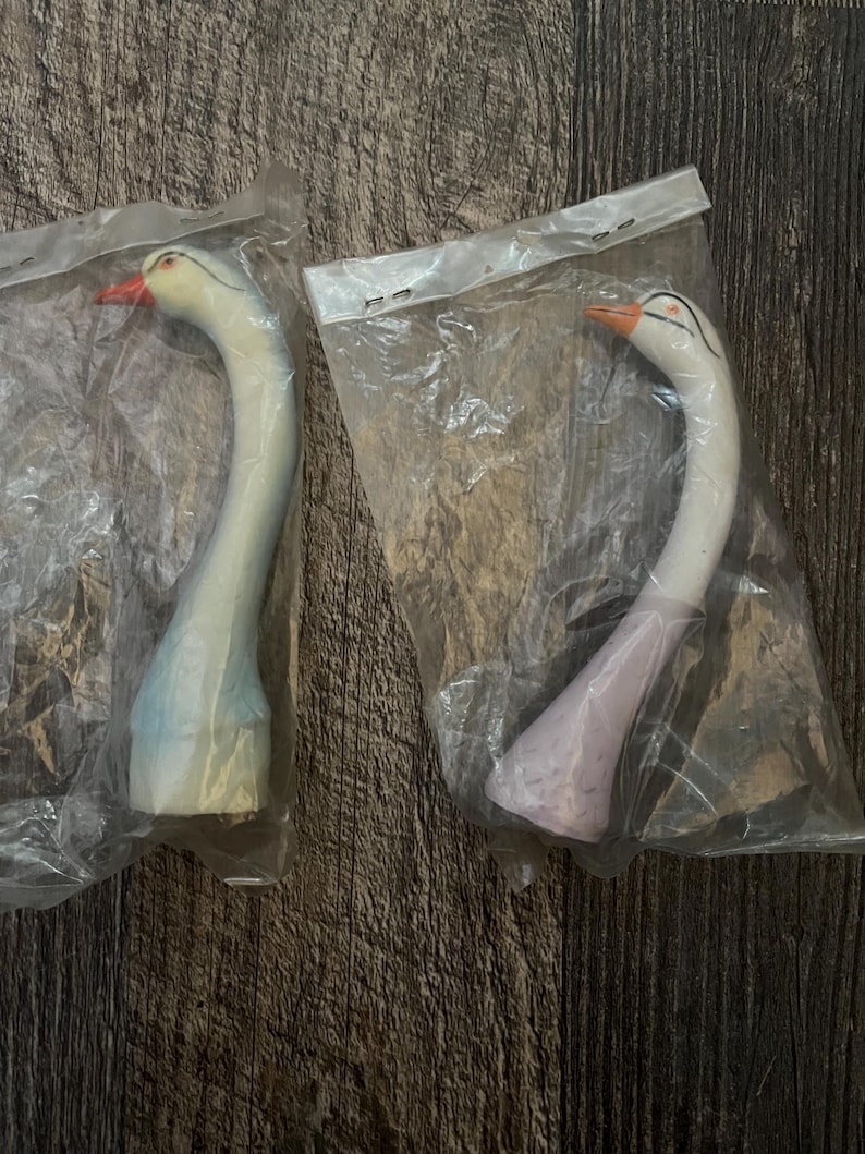 May include: Two ceramic swan figurines in individual plastic packaging. The swan on the left is light blue and white, while the swan on the right is purple and white.