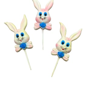 Op de afbeelding: Drie paashaas taarttoppers. Elke konijn heeft een wit gezicht met roze accenten op de oren en wangen, blauwe ogen en een rode glimlach. Een blauwe vlinderdas met roosdetail bevindt zich aan de basis van elk konijn.