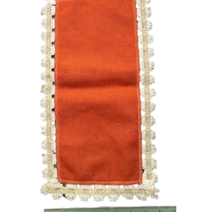 May include: An orange rectangular burlap placemat with a cream-colored, scalloped lace trim. A ruler is placed below the placemat, measuring approximately 12 inches in length.