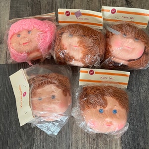 Puede incluir: Cinco cabezas de muñeca de plástico en embalaje transparente. Las cabezas de muñeca tienen diferentes colores de pelo, incluyendo rosa y marrón. Cada cabeza tiene una expresión sonriente y está etiquetada como "PLASTIC DOLL".