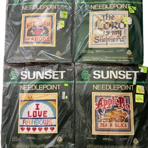 May include: Four needlepoint kits featuring colourful designs and text. The kits are packaged in clear plastic bags with black and green backgrounds. The designs include "The Kitchen is the Heart of a Home", "The Lord is My Shepherd", "I Love Rainbows", and "Apple Pie 25¢ a Slice".