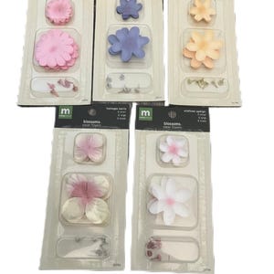 Puede incluir: Surtido de flores de papel en varios colores, incluyendo rosa, azul y amarillo. Cada paquete contiene múltiples tamaños de flores y chinchetas para sujetar. Los paquetes están etiquetados como "blossoms" y "paper flowers".