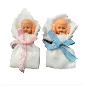 May include: Two vintage baby doll favours. Each doll is wrapped in white fabric with a pink or blue ribbon tied in a bow. The dolls have peach-coloured skin and blonde hair.