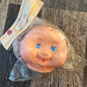 Op de afbeelding: Een plastic poppenhoofd met blauwe ogen, een roze gezicht en een kleine mond. Het hoofd is verpakt in een doorzichtige plastic zak met een label waarop "Plastic Doll" en "Made in Hong Kong" staat.