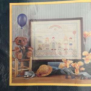May include: Counted cross stitch pattern featuring a blue and white design with the text "Grandchildren are the treasures of our lives". The pattern is framed and displayed on a table with a teddy bear, a hat, and a blue balloon.