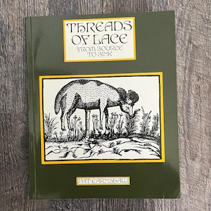 Puede incluir: Un libro titulado "Threads of Lace From Source to Sink" con una cubierta verde. La portada presenta una ilustración en blanco y negro de una oveja y texto en un recuadro amarillo. El nombre del autor, Pat Earnshaw, está en la parte inferior.