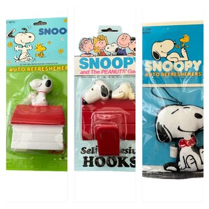 May include: Three Snoopy auto air fresheners in original packaging. One features Snoopy on his doghouse, another shows Snoopy and the Peanuts gang, and the third has a plush Snoopy with a red bow tie.