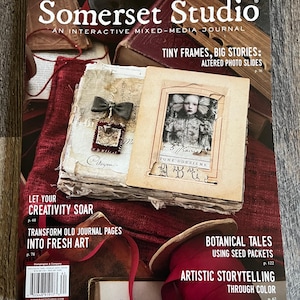 May include: The cover of "Somerset Studio" magazine, an interactive mixed-media journal. The cover features a collage of vintage ephemera, including a photograph, fabric, and text. The magazine promotes artistic storytelling and creative projects.