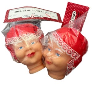 Op de afbeelding: Twee Mrs. Claus poppenhoofden in plastic verpakking. De hoofden zijn huidskleurig met blauwe ogen en rode lippen. Ze dragen rode mutsen met witte kant. De verpakking bevat de tekst "MRS. CLAUS DOLL HEAD" en "Fibre Craft".