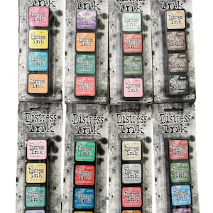 Può includere: Più confezioni di tamponi Distress Ink in vari colori. Ogni confezione contiene quattro tamponi in tonalità rosa, giallo, blu e arancione. La confezione presenta la scritta "Distress Ink" in un font invecchiato.