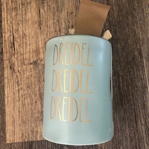 Puede incluir: Un dreidel de cerámica azul claro con letras doradas. La palabra "DREIDEL" se repite tres veces verticalmente en la parte delantera del objeto cilíndrico. Una etiqueta marrón está adjunta en la parte superior.