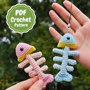 May include: Two crocheted fish keychains, one pink and one blue, with yellow accents. The keychains are hanging from a hand. The image includes the text "PDF Crochet Pattern".