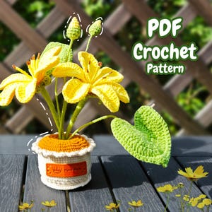May include: A crocheted flower arrangement in a white pot with an orange rim. The flowers are yellow and the leaves are green. The pot has a label that says "Hand Made". The arrangement is on a wooden table with a blurry background of a wooden fence and green foliage. The text "PDF Crochet Pattern" is in the upper right corner of the image.
