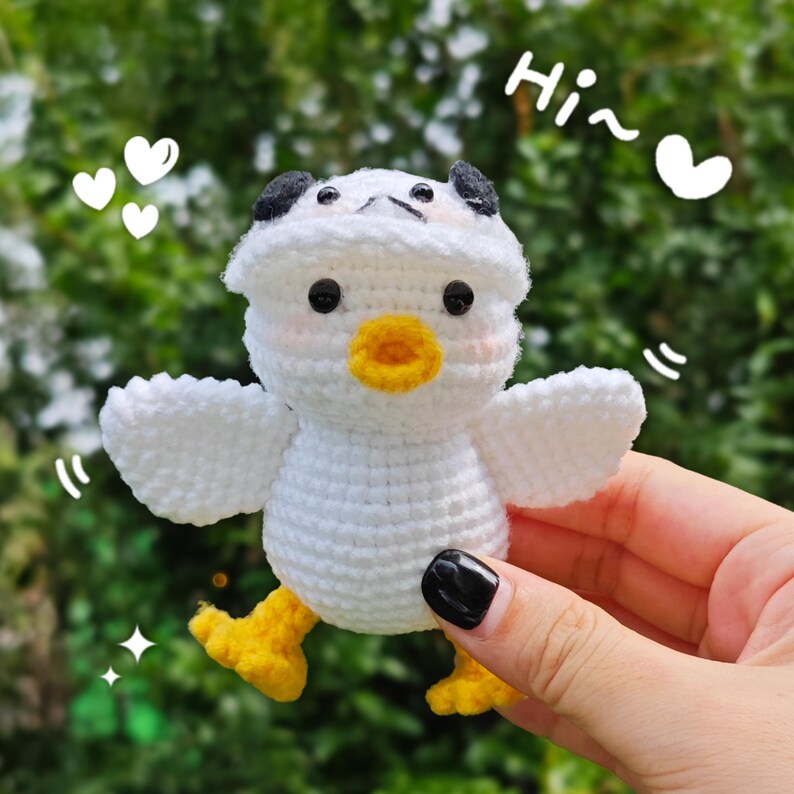 以下が含まれることがあります： 白と黒のパンダの耳と黄色いくちばしを持つ、白の編みぐるみひよこ。ひよこは羽を広げており、黄色い足をしている。背景は緑で、白い文字で「Hi~」と書かれている。