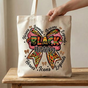 May include: A beige canvas tote bag featuring a large bow design with the words "BLACK HISTORY" in the center. The bow is decorated with colorful African patterns and the words "Heroes, Icons, Legends, Africans" are written around the bow.