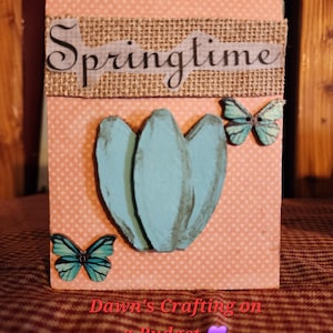 May include: A handmade spring-themed decorative block. The block features a light pink background with a polka dot pattern. It has a burlap banner with the word "Springtime" and a painted blue flower. Two butterfly accents and the text "Dawn's Crafting on a Budget" are also visible.