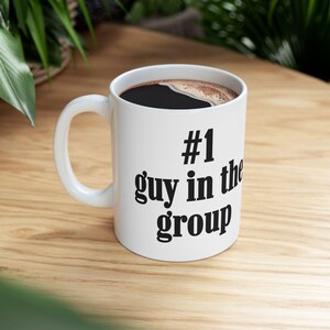 May include: White ceramic coffee mug with the text "#1 guy in the group" in black. The mug is filled with dark coffee. The mug has a white handle and sits on a wooden surface.