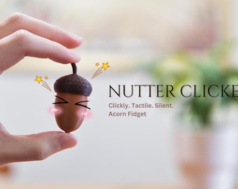 May include: A brown and dark brown acorn-shaped fidget toy held between fingers. The text "NUTTER CLICKER" is displayed, along with the words "Clickly. Tactile. Silent. Acorn Fidget."