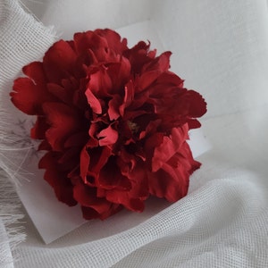 May include: A deep red artificial peony flower with numerous layered, ruffled petals. The flower is centered on a plain white square card, showcasing a rich, saturated crimson color and a dense, full-bloom structure with delicate petal edges.