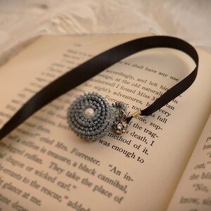 May include: A black ribbon bookmark with a blue and silver beaded charm. The charm has a pearl in the center and is attached to a silver chain. The bookmark is lying on an open book with text.