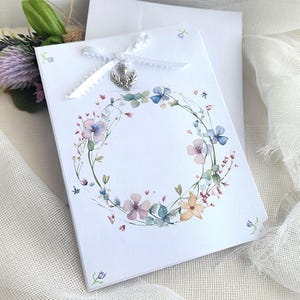 May include: A white greeting card with a floral wreath design in pastel colors. The card is adorned with a white ribbon and a silver charm. An envelope is behind the card.