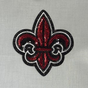 May include: A sequined fleur-de-lis patch in red, black, and silver. The design features a black outline with silver glitter, surrounding red and black sequins. The patch is on a light gray fabric.
