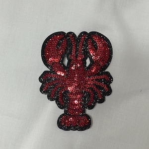 May include: A red sequined lobster patch with a black glitter outline. The patch features a detailed design of the crustacean, with its claws, body, and tail clearly defined. The sequins add a sparkling effect.