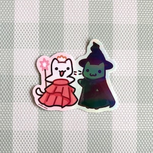 May include: A sticker featuring two cartoon cats. One cat is dressed as a fairy with a pink dress and a star wand. The other cat is dressed as a witch with a black hat and cloak. The sticker has a holographic effect.