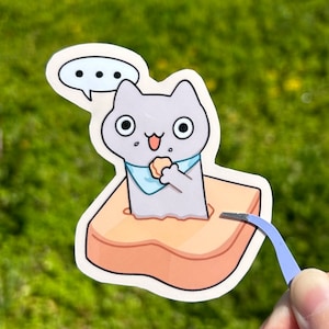 May include: A sticker depicting a cartoon cat with grey fur, large eyes, and a blue bandana, consuming a snack. The cat is inside a piece of toast, with a thought bubble above. Tweezers are in the bottom right corner.