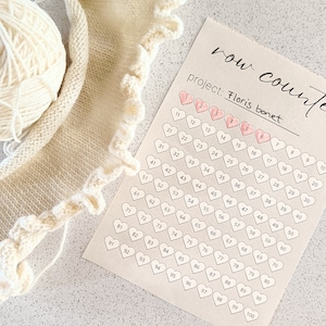 以下が含まれることがあります： ピンクのハートと「row counter」と「project: Floris bonet」のテキストが書かれた、ベージュの編み物のプロジェクトと行カウンター。
