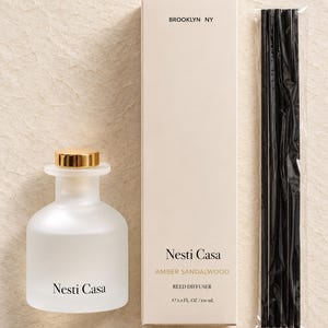 May include: A frosted glass reed diffuser bottle with a gold cap, a tall beige rectangular box labeled Nesti Casa Amber Sandalwood, and a bundle of black reeds in plastic packaging. The box text includes BROOKLYN NY and 5.0 FL. OZ / 150 mL.