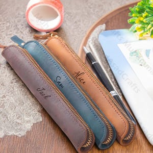 May include: Four personalized leather pen cases in shades of brown, teal, and tan, each with a gold zipper and a name: Jack, Sam, and Alice. A black pen is visible. The cases are on a wooden tray.