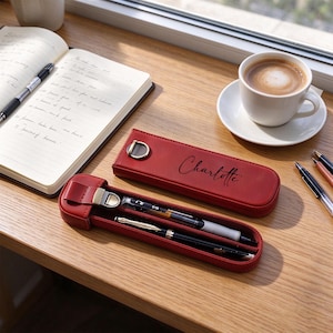 Peut inclure: Un étui à stylos en cuir rouge avec un fermoir en métal doré et le prénom Charlotte imprimé en écriture noire. L'étui est présenté fermé et ouvert, contenant deux stylos, posé sur une surface en bois près d'un carnet et d'une tasse de café.