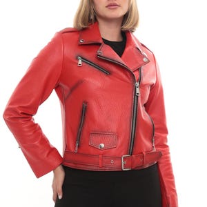 Puede incluir: Chaqueta de cuero roja de estilo motero. Presenta un cuello con solapas, cierre de cremallera asimétrico, bolsillos con cremallera y cinturón en la cintura. La chaqueta se lleva con un top negro y pantalones negros.