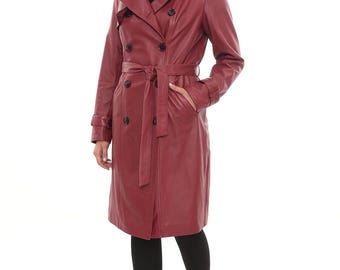 Op de afbeelding: Een bordeauxrode leren trenchcoat met een dubbelrijig ontwerp, een riem en zakken. De jas is knielang en wordt gedragen met zwarte leggings en zwarte combat boots. Het model staat voor een witte achtergrond.