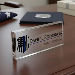 Pode incluir: Uma placa de mesa de acrílico transparente com o texto "DANIEL RODRIGUEZ POLICE SERGEANT". A placa apresenta um gráfico de distintivo de polícia preto e azul. A placa está em uma mesa de madeira marrom escura.