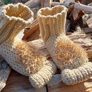 May include: A pair of beige crocheted slippers with fuzzy brown trim. The slippers are sitting on a pile of wood.