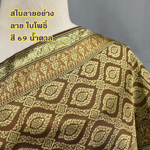 Pode incluir: Close-up de um tecido marrom e dourado estampado. O tecido tem um desenho floral repetido com uma borda geométrica. O texto em tailandês é visível no canto superior esquerdo. O tecido está drapeado sobre um manequim.