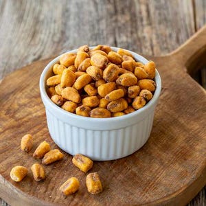 May include: A white, ridged ceramic bowl filled with roasted corn kernels sits on a wooden cutting board. The corn is golden brown and scattered around the bowl. The cutting board rests on a weathered wooden surface.