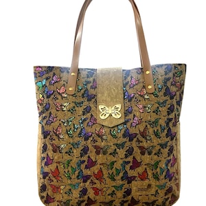 May include: A cork tote bag with a butterfly print. The bag has brown leather straps and a gold butterfly clasp. The bag is made by the brand "ThinkCork."