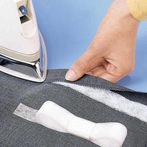 May include: A white iron being used on a dark gray fabric with a white strip of fusible interfacing. The iron is positioned near the edge of the fabric, and a hand is holding the fabric in place. The background is a solid light blue.