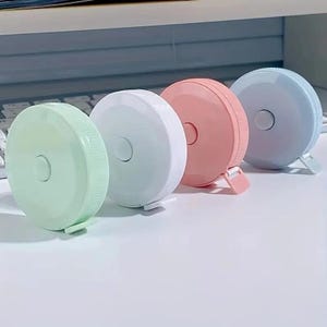 May include: Four colorful retractable tape measures. The tape measures are in shades of light blue, pink, white, and green. Each tape measure has a round, flat case with a button in the center.