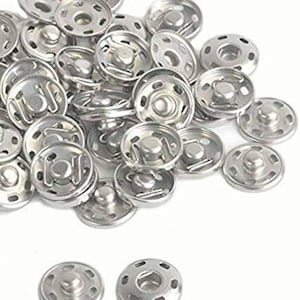May include: A pile of silver metal snap fasteners, also known as poppers, for sewing projects. The fasteners are round and have a raised centre with a small hole in the middle.