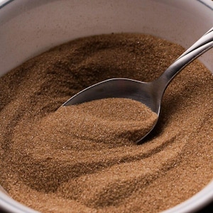 Op de afbeelding: Een witte kom gevuld met bruine kristalsuiker met een zilveren lepel die gedeeltelijk in de suiker is ondergedompeld.