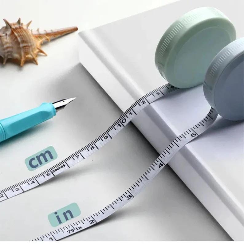 May include: Two measuring tapes with white tape and black markings. The tape measures in inches and centimeters. The tape is wound around a light blue and a light gray spool.