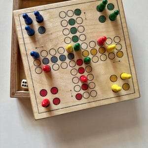 Op de afbeelding: Een houten bordspel met een lichtbruine afwerking. Het spelbord heeft een kruisvormig ontwerp met gekleurde cirkels en pionnen in blauw, groen, rood en geel. Een houten doos bevat het bord en een witte dobbelsteen. Een klassiek familiespel.