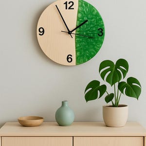 Könnte beinhalten: Eine Uhr aus Naturholz und Grün mit schwarzen Zahlen und Zeigern. Die Uhr hängt an einer hellgrauen Wand mit einer grünen Pflanze in einem beigefarbenen Topf und einer hellblauen Vase auf einem hellen Holzregal.