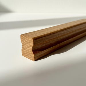 May include: A section of unfinished, light brown wooden molding with a decorative edge. The wood grain is visible, and the piece is rectangular with a curved profile on one side. The molding is likely for interior design or carpentry projects.