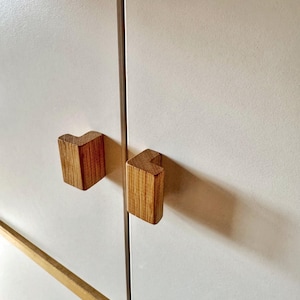 Pode incluir: Close-up de uma porta de armário branca com duas puxadores de madeira em forma de L. Os puxadores são feitos de madeira clara e são montados na porta do armário. Uma fina haste de metal corre verticalmente entre os puxadores.