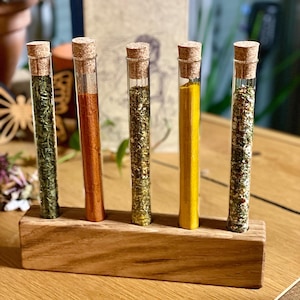 Peut inclure: Cinq tubes à essai en verre avec des bouchons en liège remplis de différentes herbes et épices séchées. Les tubes à essai sont disposés dans un support en bois.